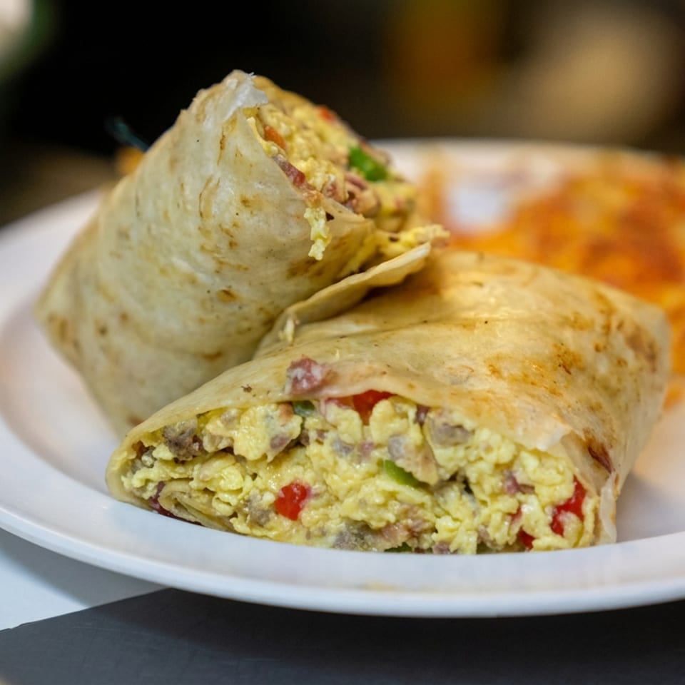 Two breakfast burritos filled with scrambled eggs, vegetables, and meat sit on a white plate with orange salsa.