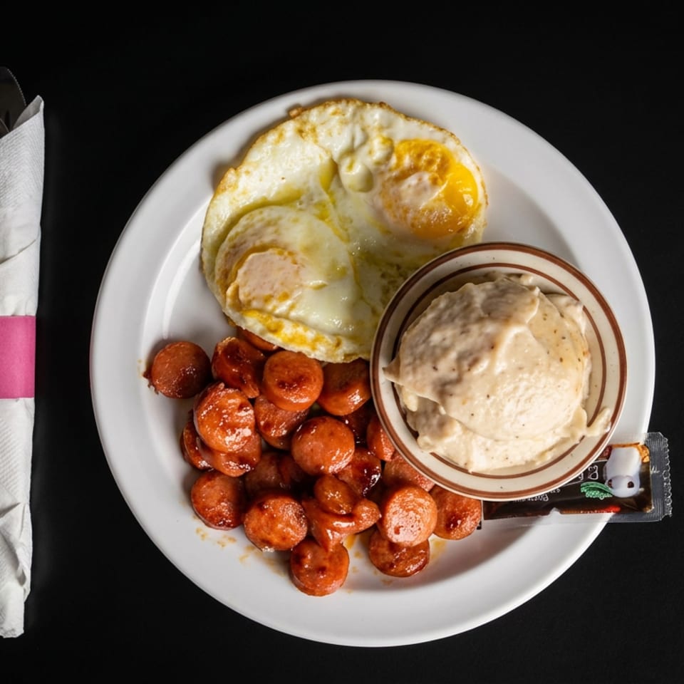 Plate of fried eggs, sliced sausage, and creamy dipping sauce with wrapped utensils on a black table.