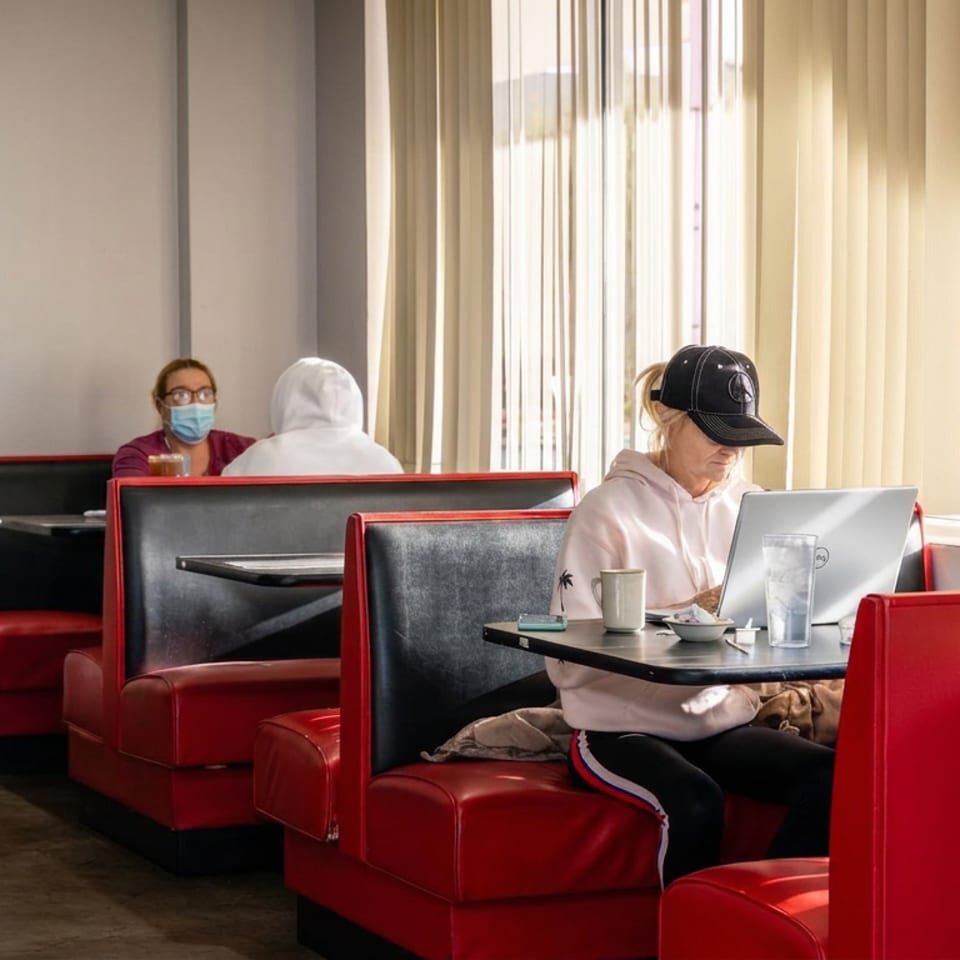 Diner interior with red booths, a masked patron, and a person working on a laptop beside sunlit windows.