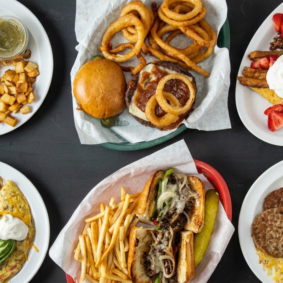Overhead spread of diner dishes including burgers, fries, onion rings, French toast, breakfast platters, and hash browns on a dark table.