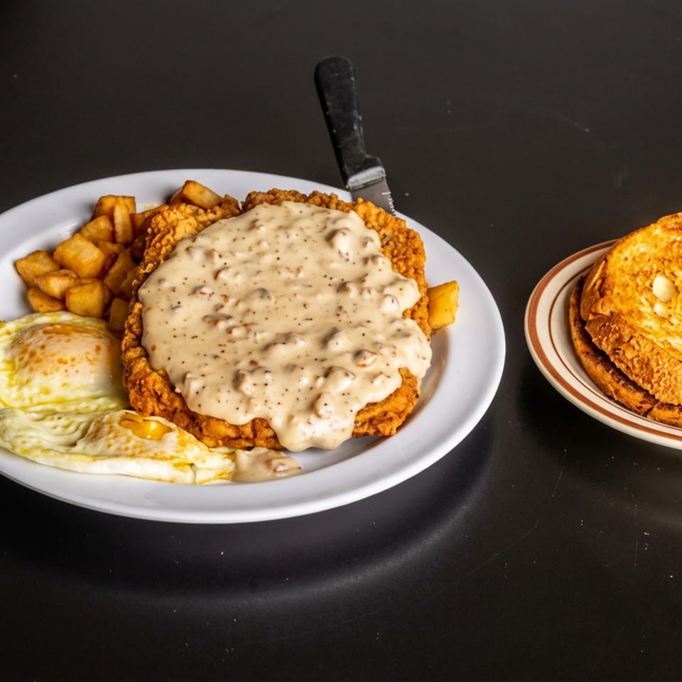 Country-fried steak topped with creamy gravy, eggs and potatoes, served with grilled cheese sandwich on a dark table.