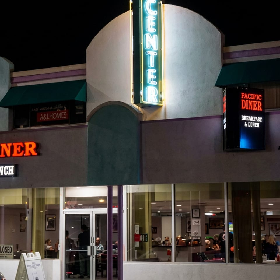 Pacific Diner storefront at night, featuring illuminated signage, large windows, and diners visible inside.